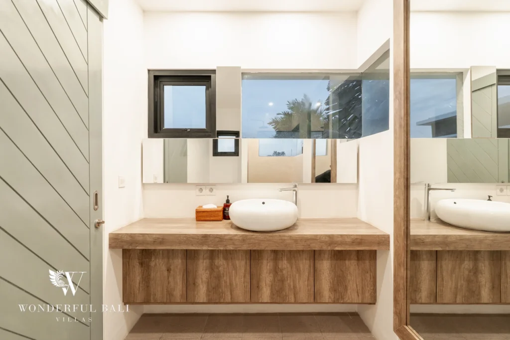A bathroom with a wooden floating vanity, single vessel basin, mirrors and windows overlooking palm trees beside a slatted door at a 3-bedroom villa in Berawa, Bali