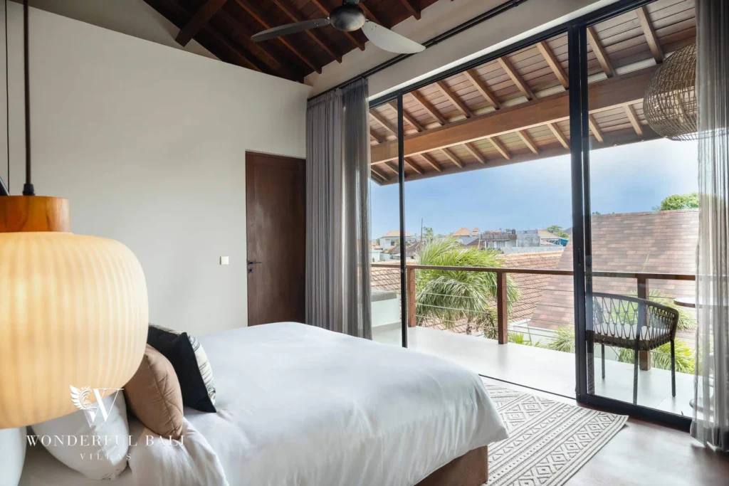 An upstairs bedroom with a king bed and sliding doors opening to a balcony with a chair and rooftop views at a 2-bedroom villa in Canggu, Bali