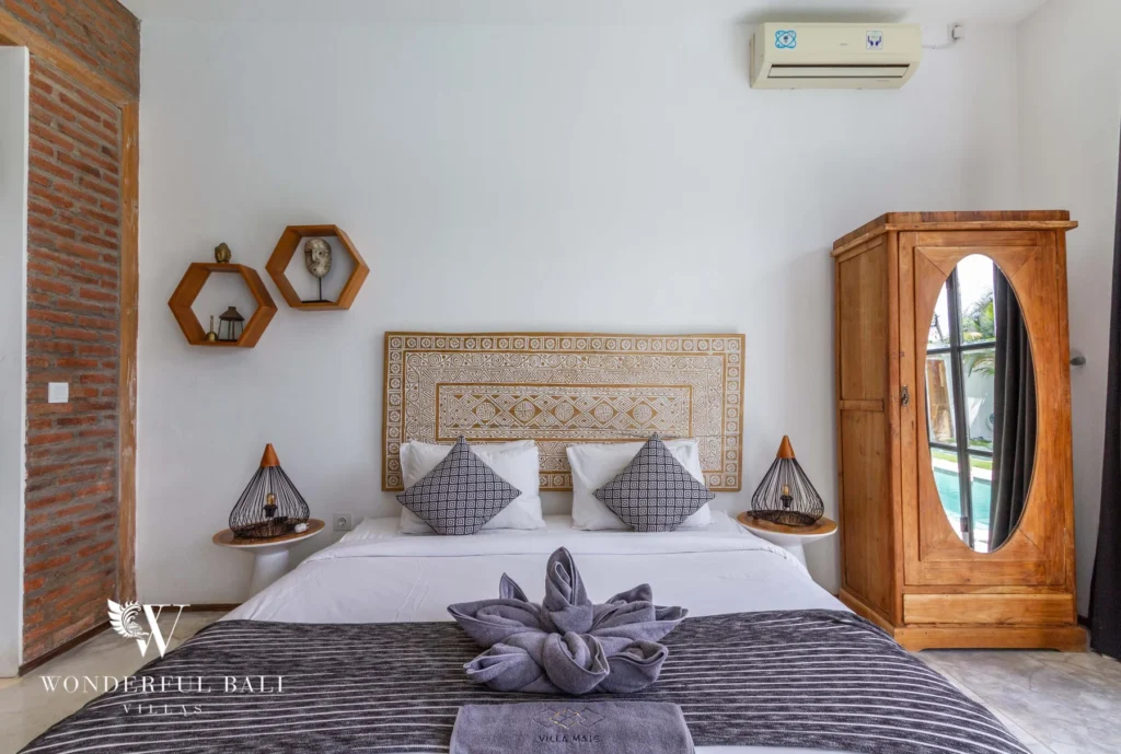 A bedroom with an ornate carved headboard, hexagonal wall shelves, brick accent wall and a mirrored timber wardrobe at a 3-bedroom villa in Seminyak, Bali