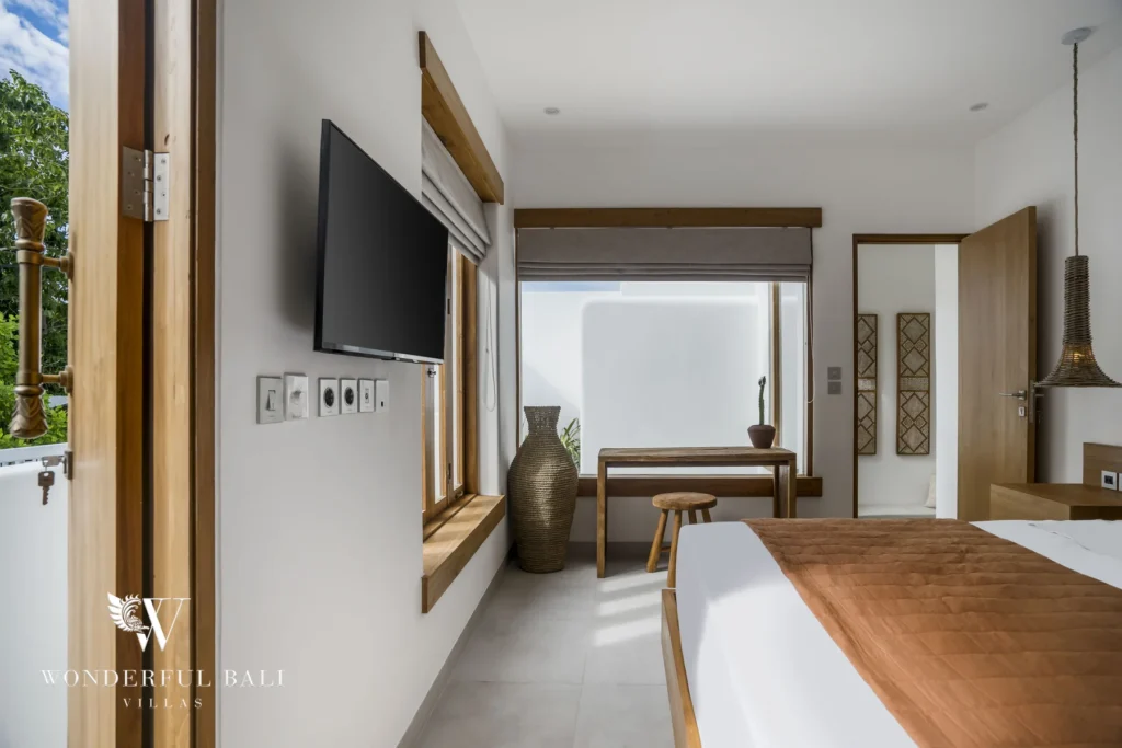 A bedroom corner with a wall-mounted TV, a slim wooden desk and stool by the window and a doorway leading toward the en-suite at a 3-bedroom villa in Bingin, Uluwatu, Bali
