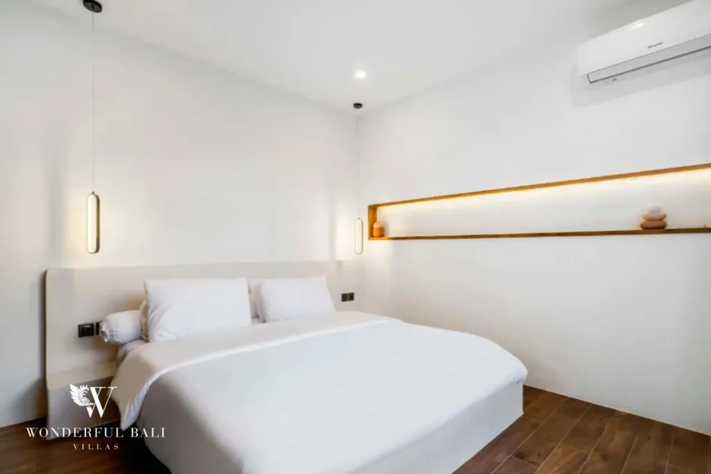 A minimalist bedroom with white bedding, a floating wooden shelf, and pendant lights at a 3-bedroom villa in Pererenan, Bali