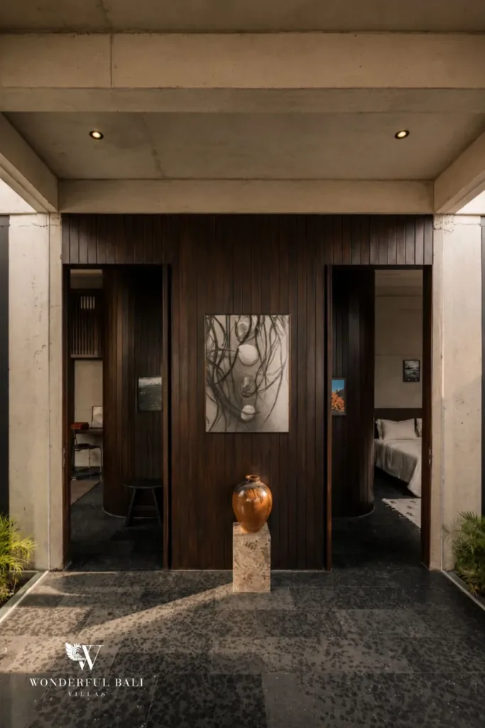 Entrance vestibule with dark timber walls, a copper vase on a stone pedestal and glimpses into two bedrooms on either side at a 2-bedroom villa in Seseh, Bali