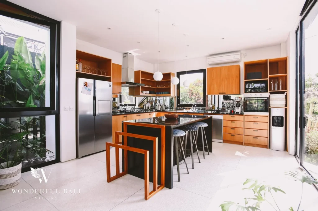 A fully equipped kitchen with a central island, bar stools, espresso machine and timber cabinetry beside a garden window at a 4-bedroom villa in Batu Bolong, Canggu, Bali
