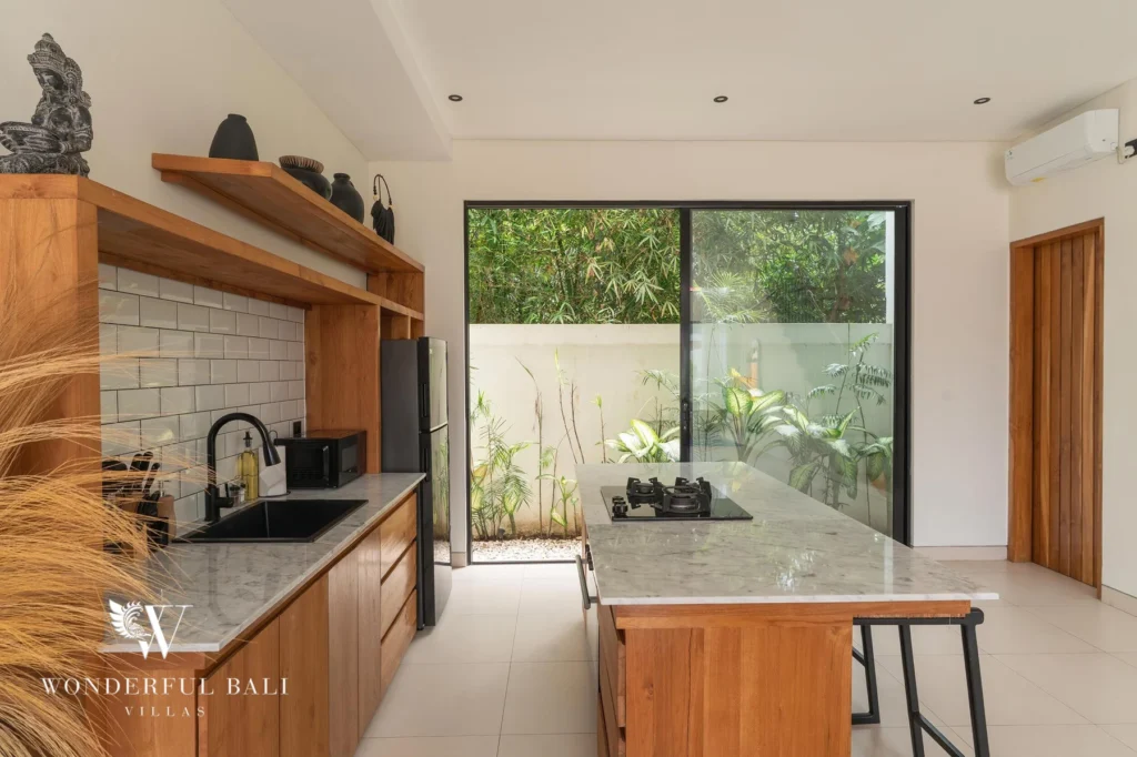 A timber kitchen with marble counters and gas hob opening through glass doors to a planted courtyard at a 2-bedroom villa in Canggu, Bali