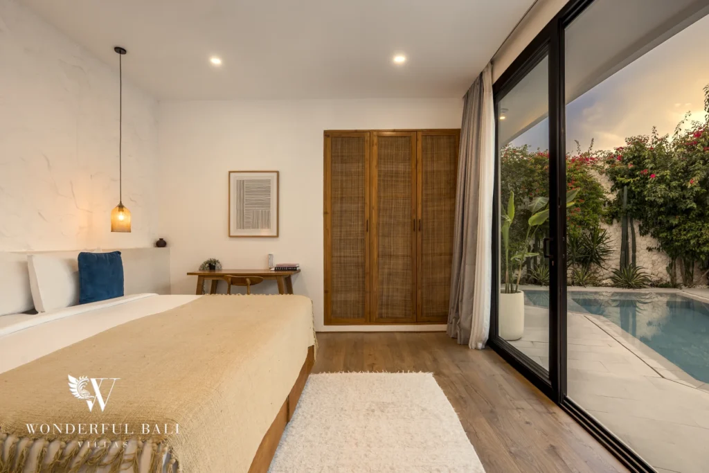 Open-plan living and dining space with a grey sofa, wooden dining table and kitchen opening onto the pool at dusk at a 3-bedroom villa in Bingin, Uluwatu, Bali