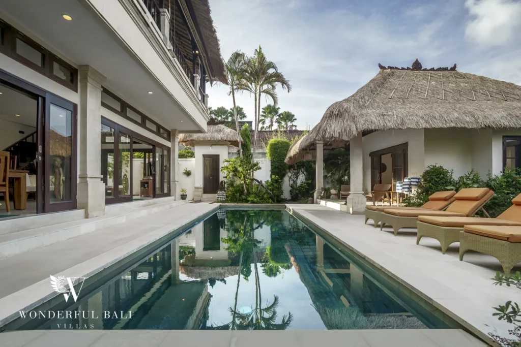 A swimming pool reflecting palm trees, edged by cushioned loungers and thatched pavilions at a 4-bedroom villa in Seminyak, Bali