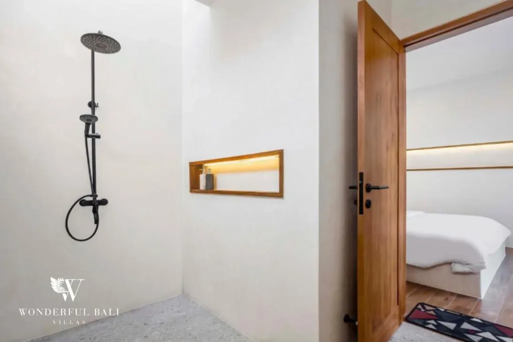 A bathroom with a black rainfall shower, micro-cement walls, and a timber-framed door leading to a bedroom at a 3-bedroom villa in Pererenan, Bali