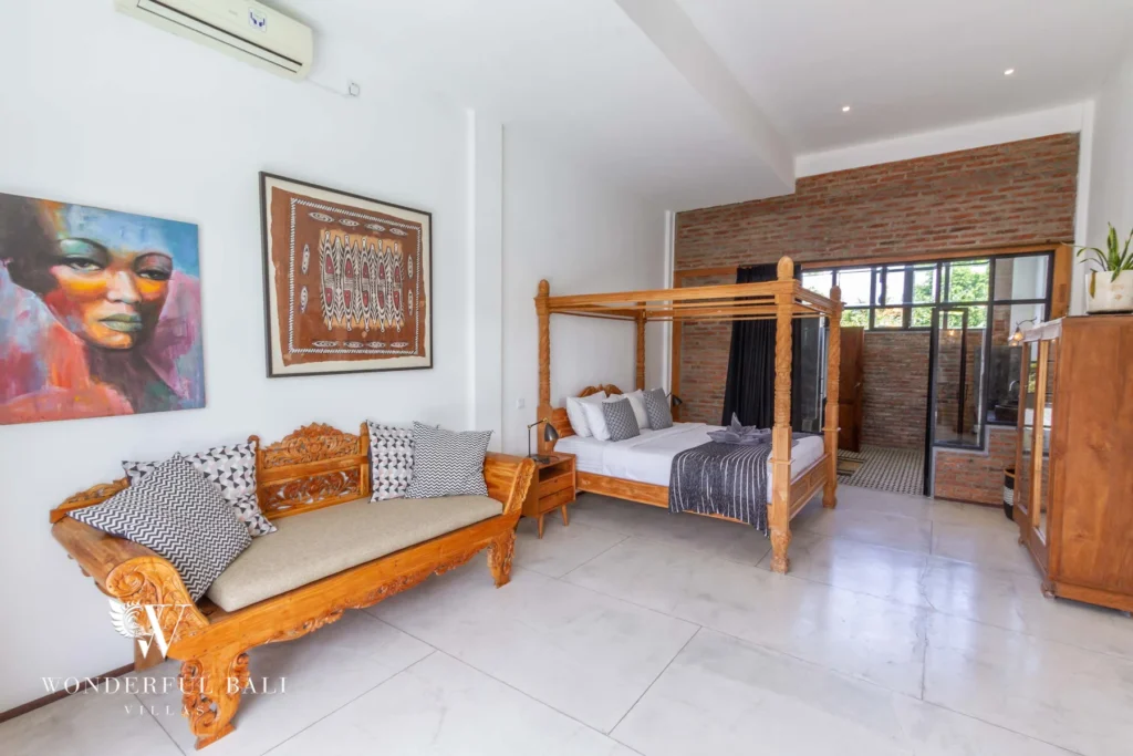A spacious bedroom with a carved four-poster bed, a wooden daybed, colourful artworks and a brick wall with an en-suite shower area at a 3-bedroom villa in Seminyak, Bali