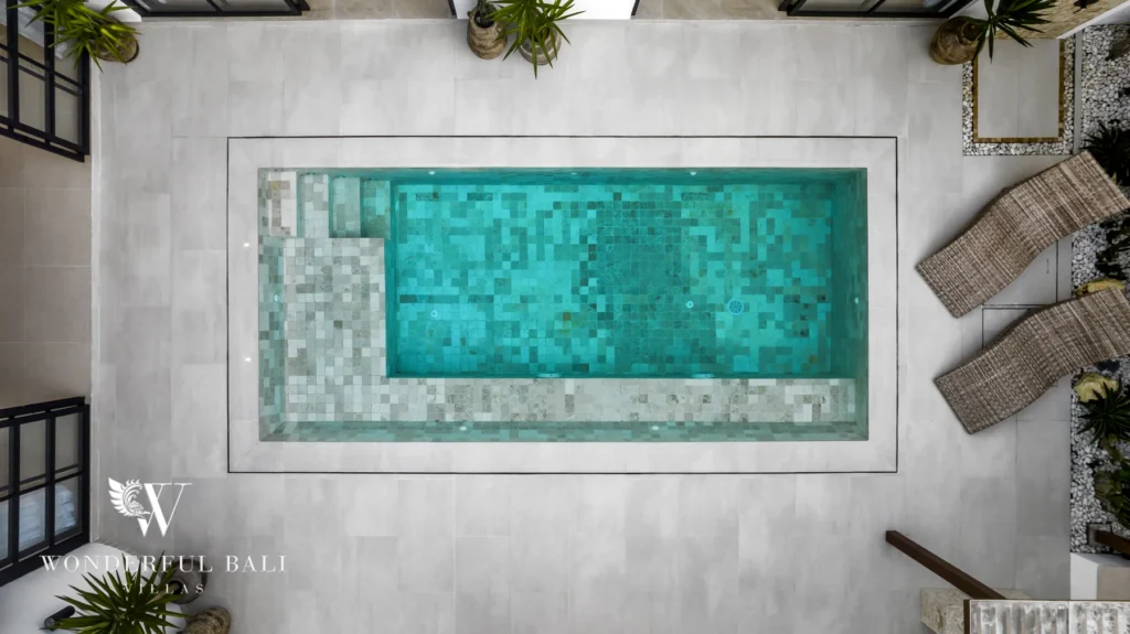 An overhead drone aerial view of the private pool with mosaic tiling flanked by rattan loungers and potted plants at a 3-bedroom villa in Bingin, Uluwatu, Bali