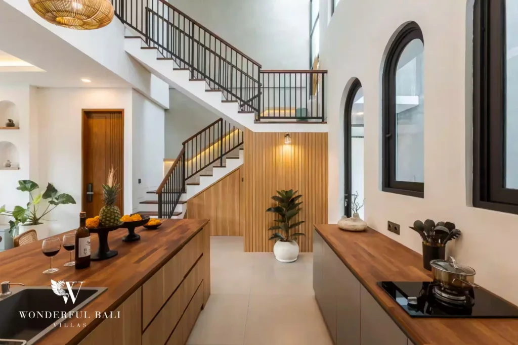 A galley kitchen with timber-topped counters, induction cooktop and staircase rising beneath a double-height window at a 4-bedroom villa in Pererenan, Bali
