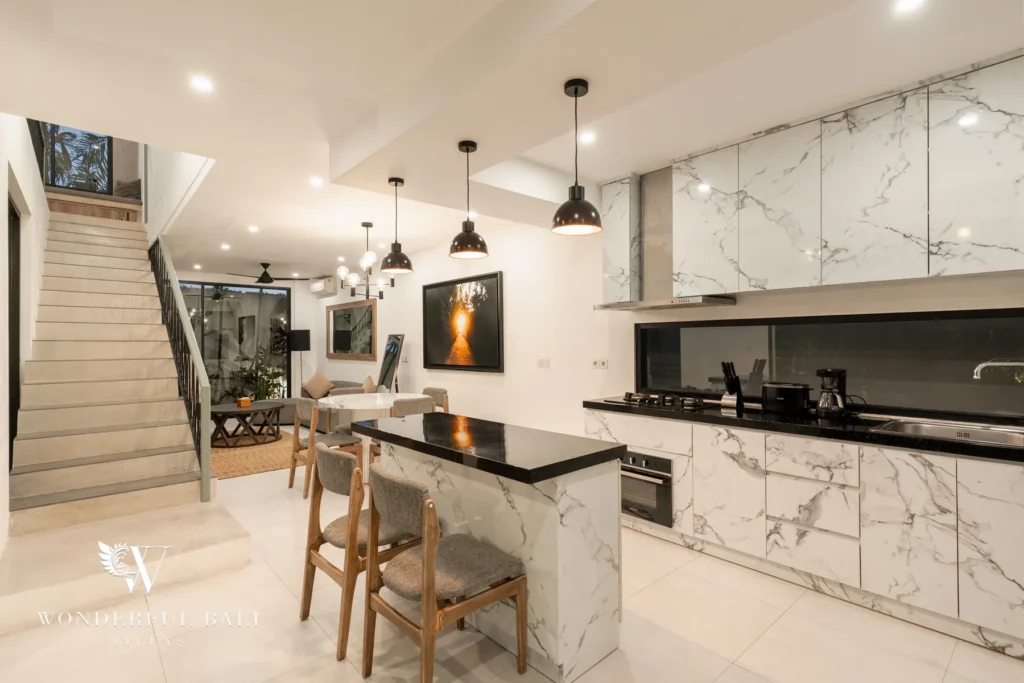 A marble kitchen with a black-topped island bar, gas hob, pendant lights and a staircase leading to the adjoining dining and lounge space at a 3-bedroom villa in Berawa, Bali