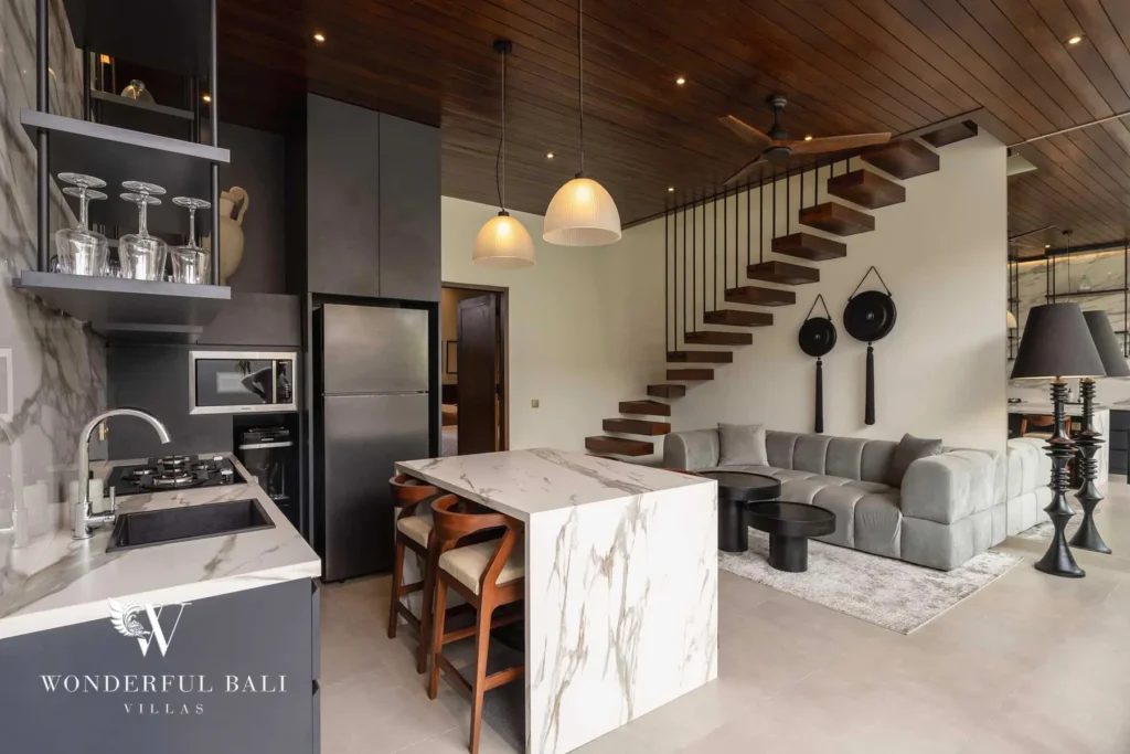 A marble kitchen and island opening to a grey sofa lounge with a floating staircase and wall gongs at a 2-bedroom villa in Canggu, Bali