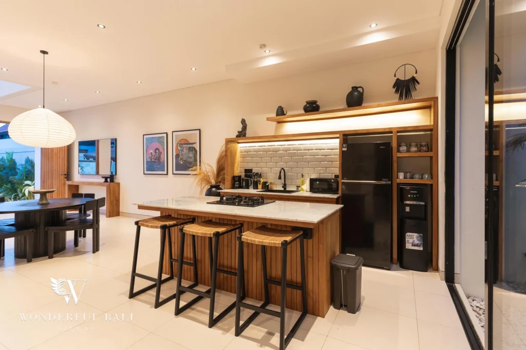 A warmly lit kitchen at night with a marble island, rattan bar stools and adjoining dining area at a 2-bedroom villa in Canggu, Bali