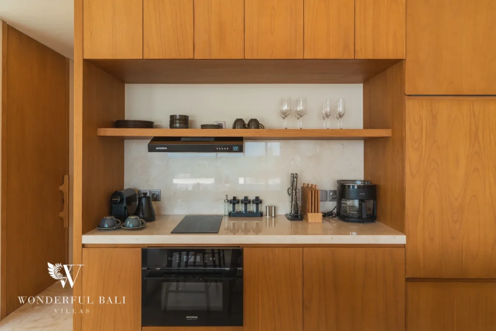 A compact timber kitchenette with an oven, cooktop, coffee machine and open shelving with glassware at a 3-bedroom villa in Bingin, Uluwatu, Bali