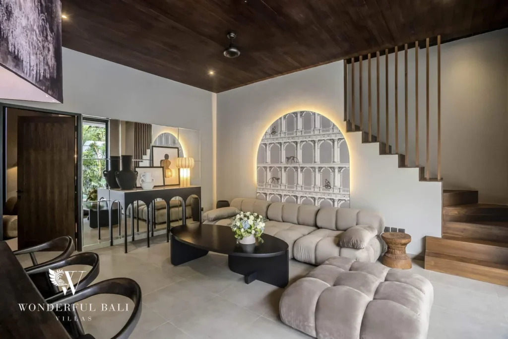 A lounge with modular velvet sofas, a black coffee table, an arched backlit wall panel and a wooden staircase under a dark ceiling at a 3-bedroom villa in Pererenan, Bali