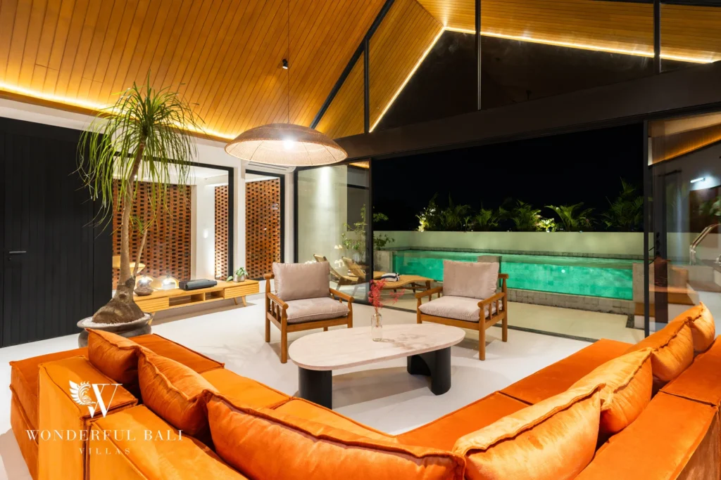 An enclosed living lounge at night with burnt-orange sofas and timber armchairs under a warmly lit vaulted ceiling, opening to an illuminated rooftop pool at a 5-bedroom villa in Batu Bolong, Canggu, Bali
