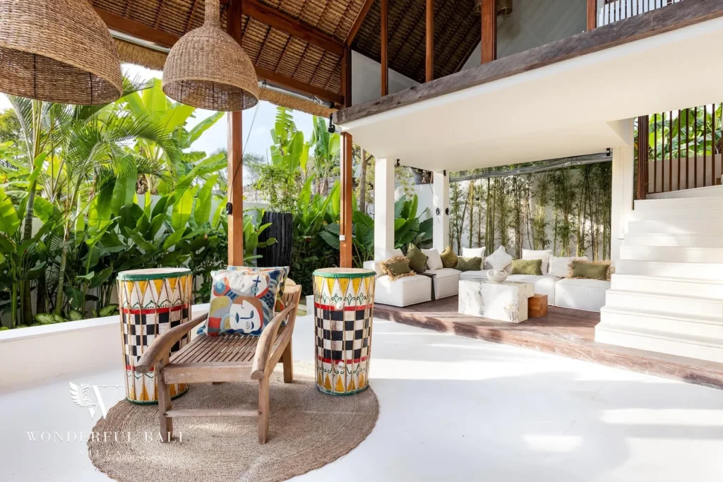 An open living pavilion with a white sofa, wooden chairs, painted drum stools and a staircase looks out over the tropical garden at a 4-bedroom villa in Canggu, Bali