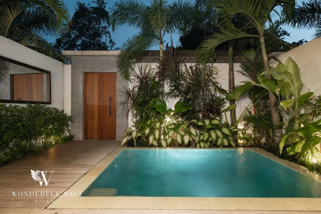 2BR villa for rent in Pererenan — pool, a private pool glowing at dusk beside a timber deck and a lush screen of palms and tropical plants by a wooden door at a 2-bedroom villa