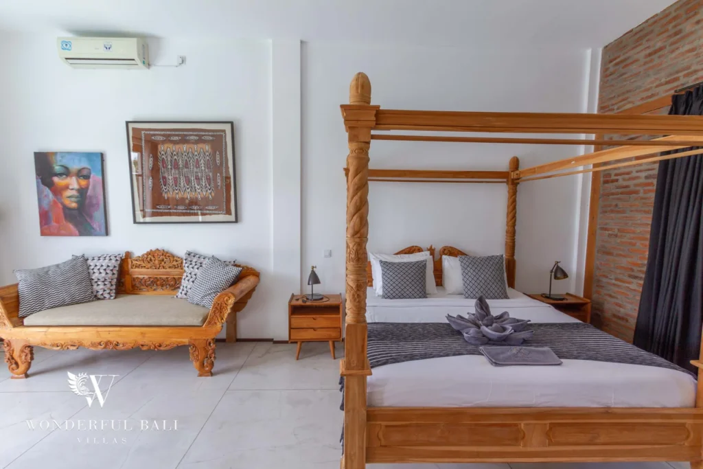 A four-poster bed beside a carved wooden daybed, framed textile art and a brick accent wall with an in-room shower at a 3-bedroom villa in Seminyak, Bali