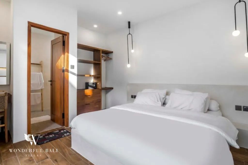 A bedroom with white bedding, wooden built-in shelving, and an ensuite doorway lit by warm sunlight at a 3-bedroom villa in Pererenan, Bali