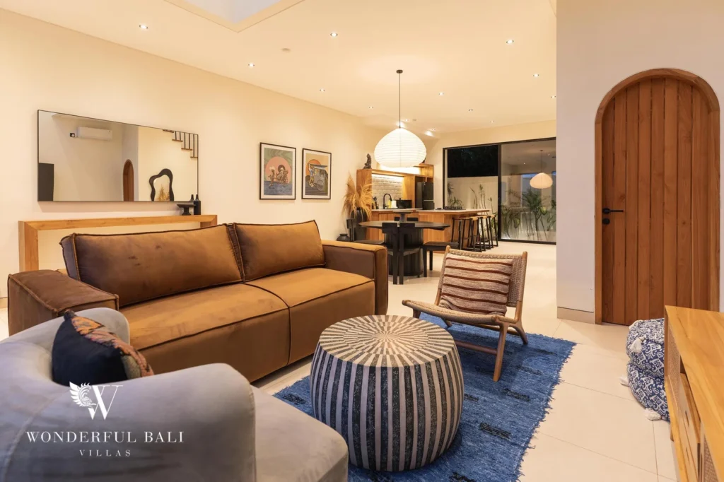 A living room at night with a brown sofa, striped pouffe and blue rug beside the dining space and bar at a 2-bedroom villa in Canggu, Bali