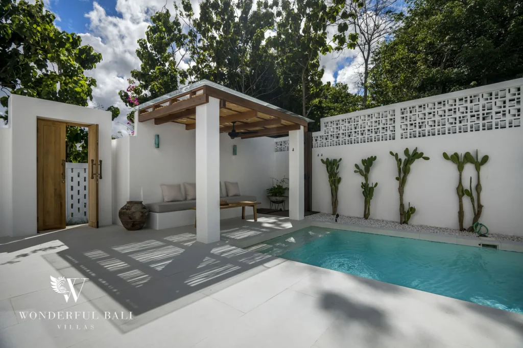 A covered outdoor lounge with a built-in cushioned sofa and low wooden table beside a plunge pool, cacti and a carved timber entry door at a 3-bedroom villa in Bingin, Uluwatu, Bali