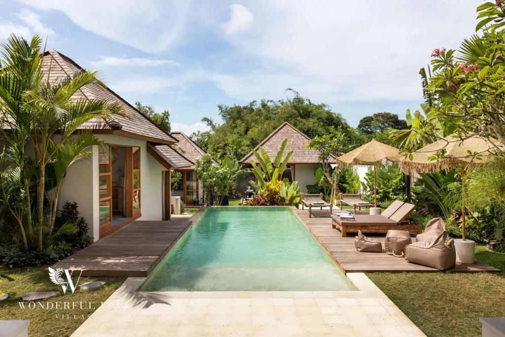 A private pool bordered by wood decking, sun loungers and bean bags runs between shingle-roofed garden bungalows under a blue sky at a 4-bedroom villa in Canggu, Bali