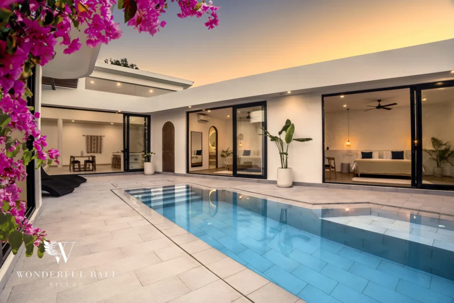 Sunset hero shot of the private pool glowing under a golden sky with illuminated bedrooms behind sliding glass doors and pink bougainvillea at a 3-bedroom villa in Bingin, Uluwatu, Bali