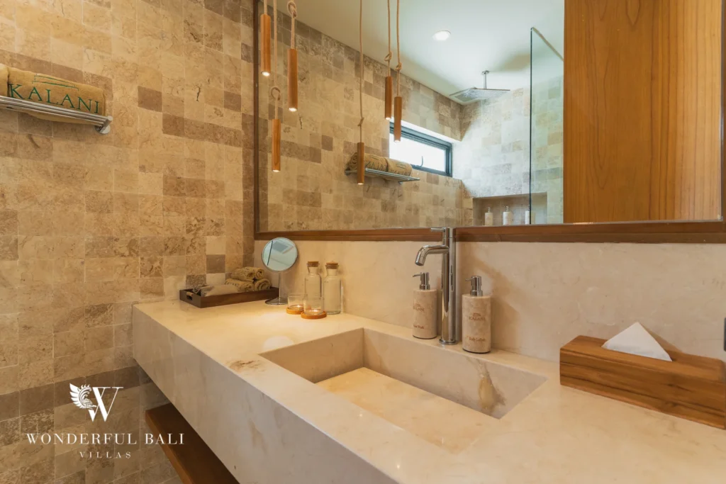 A bathroom with a stone-tiled wall, a large mirror, hanging pendant lights and a carved stone sink at a 3-bedroom villa in Bingin, Uluwatu, Bali