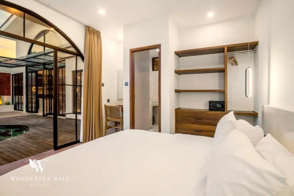 A bedroom with built-in timber shelving and arched steel-frame doors opening onto the pool deck at sunset at a 3-bedroom villa in Pererenan, Bali