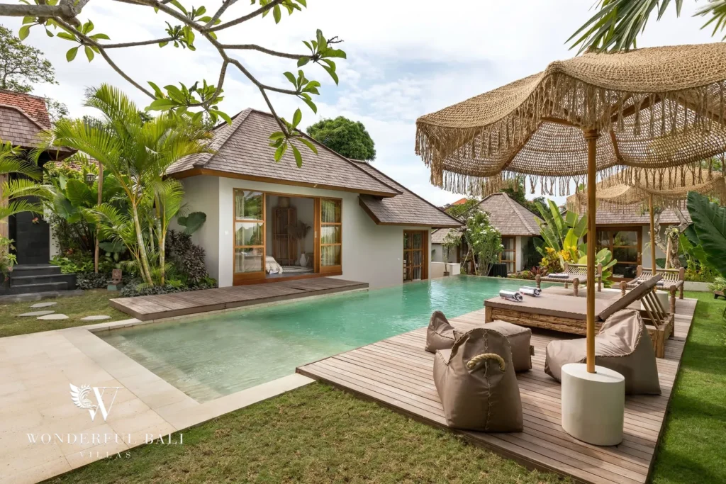 A private pool with a fringed umbrella, sun loungers and bean bags on wood decking sits beside a garden bungalow with sliding doors at a 4-bedroom villa in Canggu, Bali