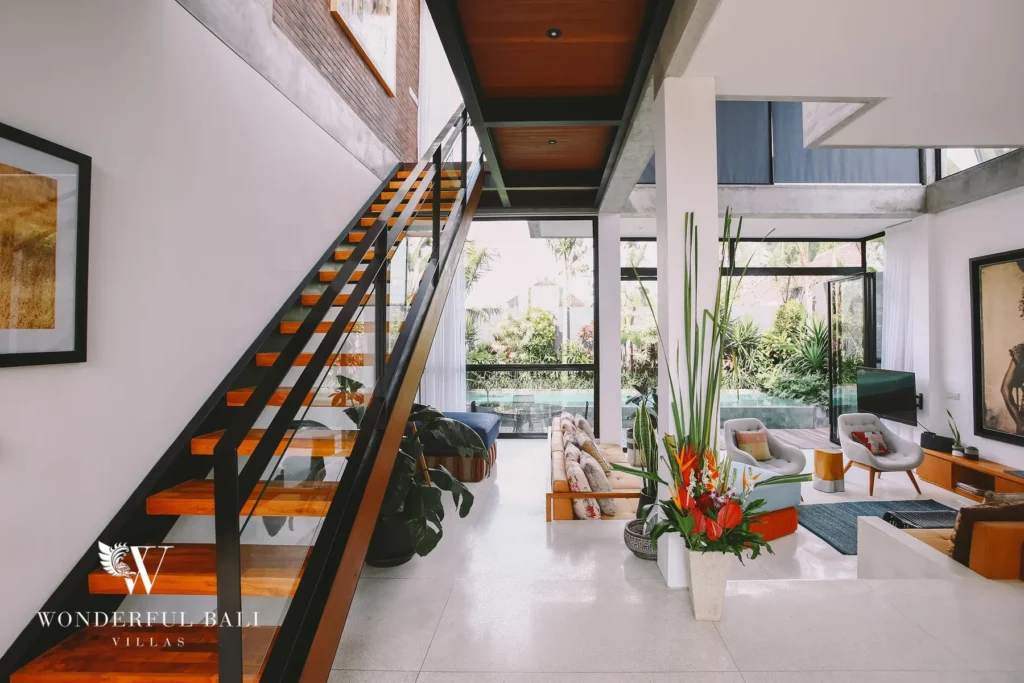 A floating timber-and-steel staircase beside white walls with a lounge area and glass doors onto the garden at a 4-bedroom villa in Batu Bolong, Canggu, Bali