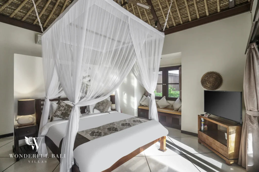 A canopy bed with white netting facing a television and a window daybed under a thatched ceiling at a 4-bedroom villa in Seminyak, Bali
