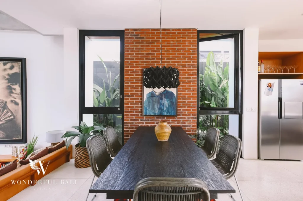 A long dark timber dining table with woven chairs set against a red-brick feature wall and black pendant light at a 4-bedroom villa in Batu Bolong, Canggu, Bali