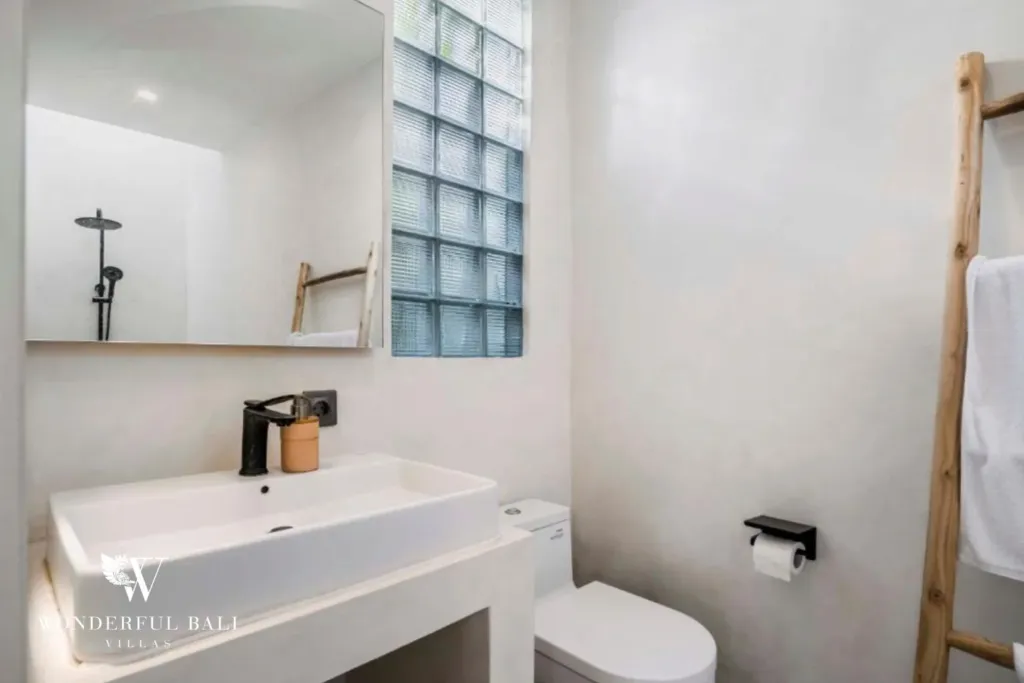 An ensuite bathroom with a white vessel sink, large mirror, glass-block window, and timber ladder towel rail at a 3-bedroom villa in Pererenan, Bali