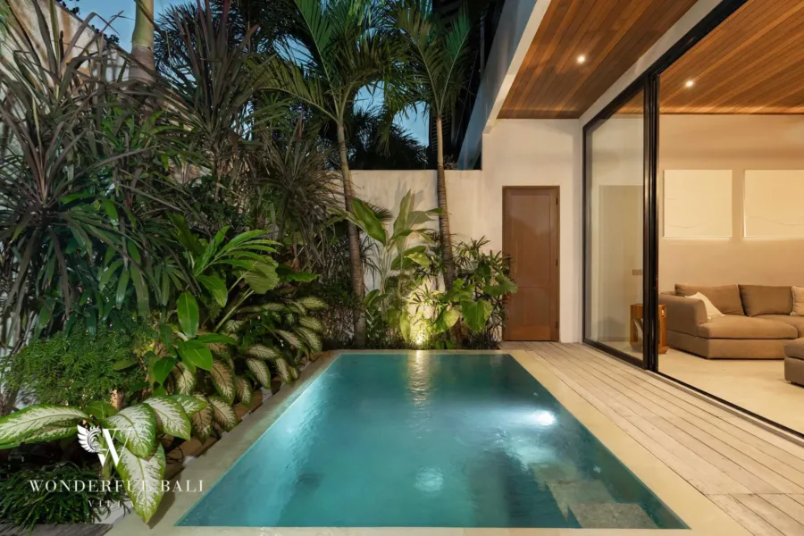 2BR villa for rent in Pererenan — pool, a private pool illuminated at dusk amid dense tropical planting beside a glass-walled lounge under a timber ceiling at a 2-bedroom villa