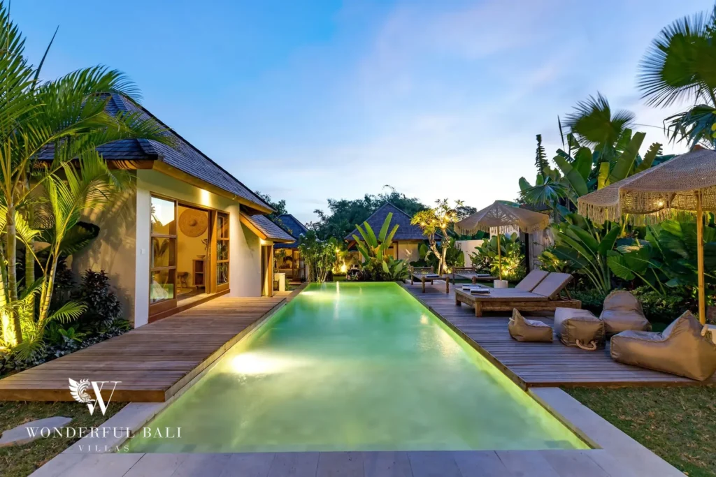 At dusk the illuminated pool glows turquoise beside wood decking with sun loungers and bean bags among tropical plants and bungalows at a 4-bedroom villa in Canggu, Bali
