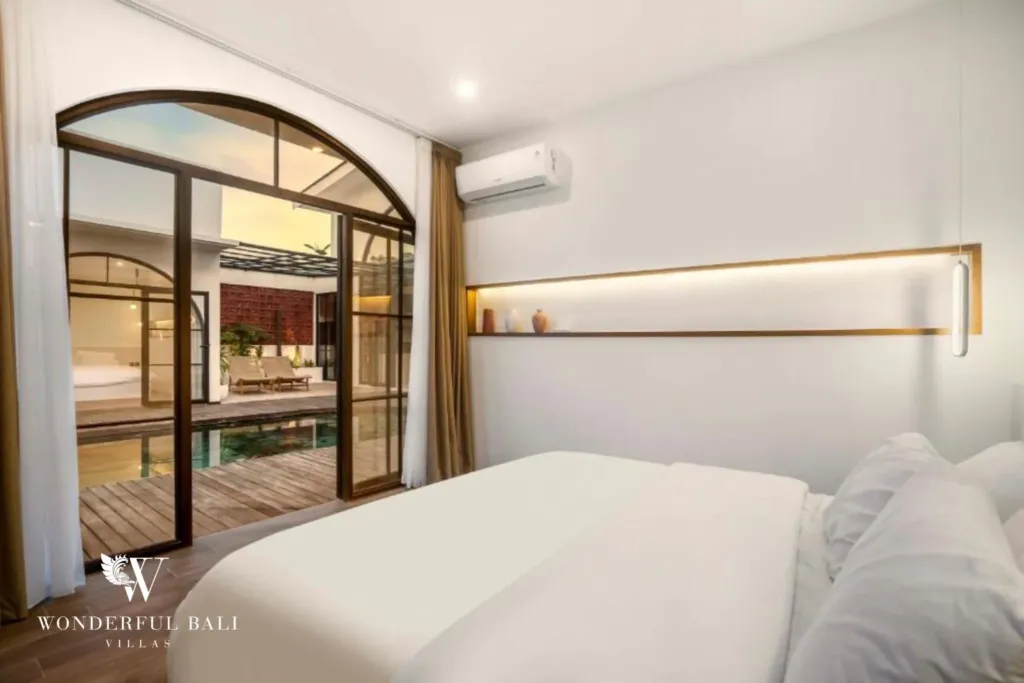 A bedroom with white bedding and a warm-lit shelf beside arched glass doors framing the pool and deck at a 3-bedroom villa in Pererenan, Bali