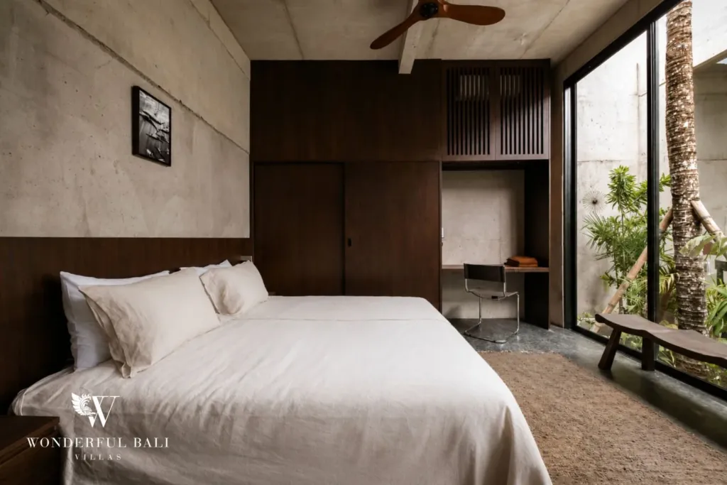 Second bedroom with a white-linen bed, timber wall panels, a built-in desk workspace and glass onto a green courtyard at a 2-bedroom villa in Seseh, Bali