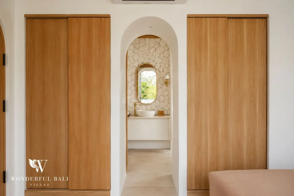 A corridor between wooden wardrobes framing an arched view of a stone-walled bathroom vanity, at a 2-bedroom villa in Bingin, Uluwatu, Bali