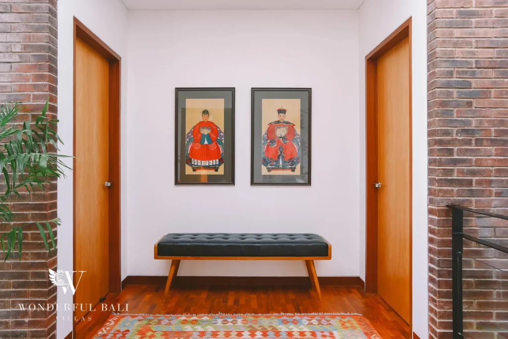 A hallway with exposed red-brick columns, timber doors, a leather bench and framed portrait artwork over a patterned rug at a 4-bedroom villa in Batu Bolong, Canggu, Bali