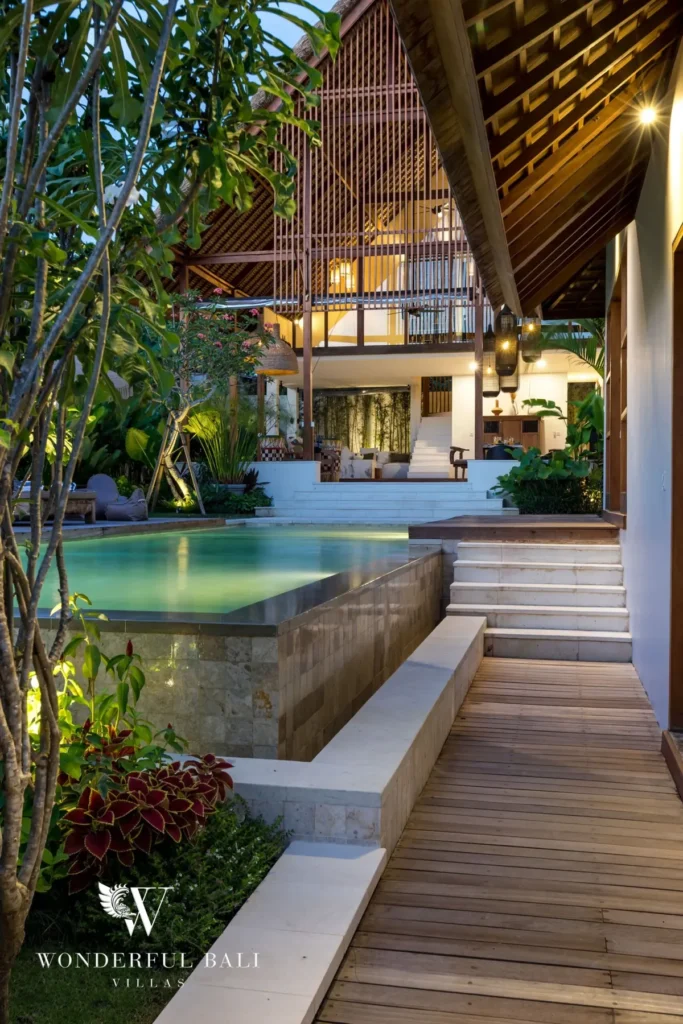 At twilight a raised pool with stone-clad sides glows beside a wooden walkway leading to the lit two-storey pavilion at a 4-bedroom villa in Canggu, Bali