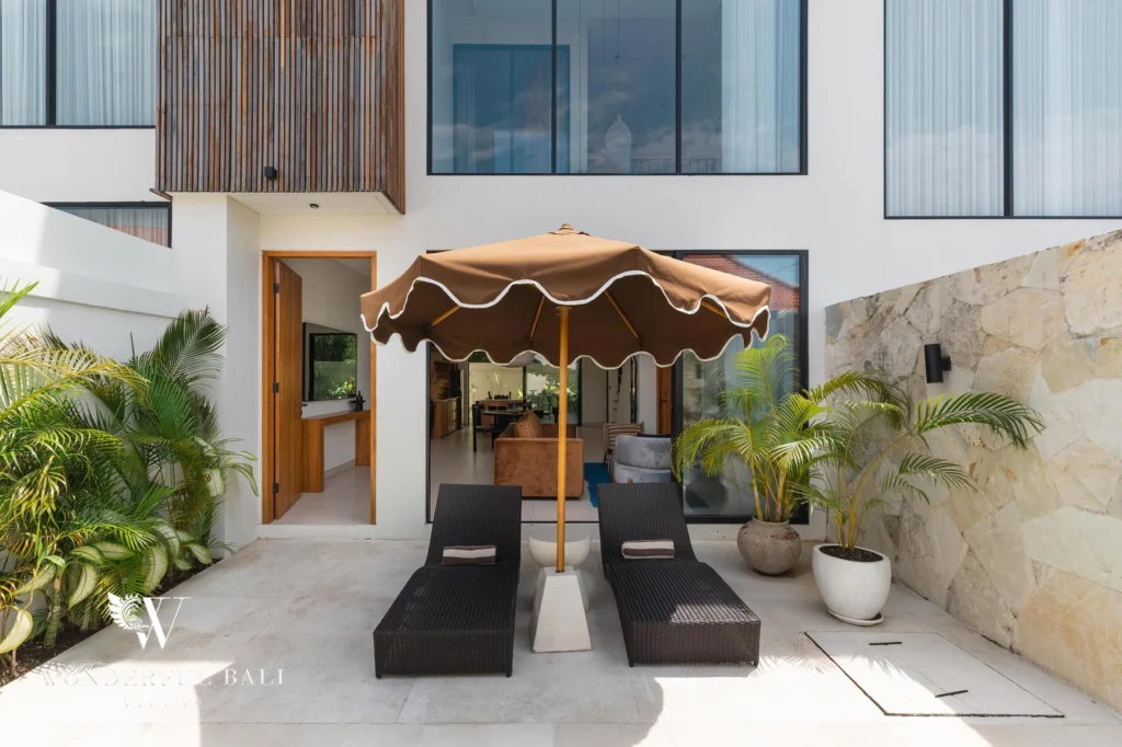 A paved poolside terrace with black sun loungers under a scalloped parasol and open doors to the living area at a 2-bedroom villa in Canggu, Bali