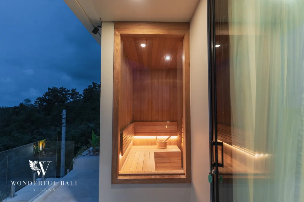 A cedar-lined sauna glowing at dusk beside a glass-railed terrace overlooking the treetops at a 3-bedroom villa in Bingin, Uluwatu, Bali