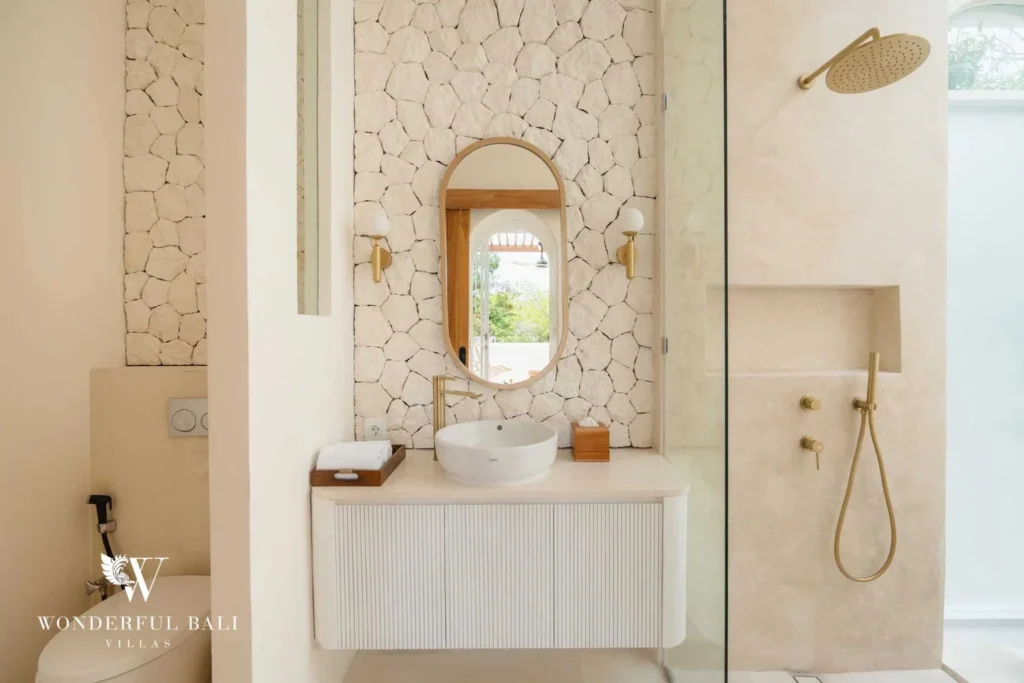 A bathroom with a stone-clad wall, oval mirror, fluted vanity and a glass walk-in shower with a gold rain head, at a 2-bedroom villa in Bingin, Uluwatu, Bali
