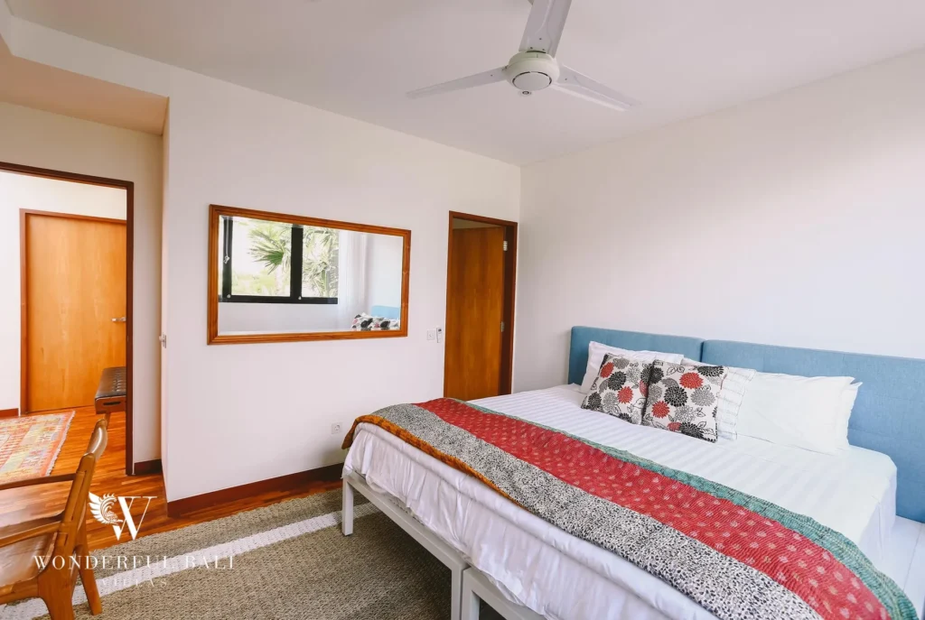 A bedroom with a blue headboard, patterned throw, ceiling fan and a wall mirror beside a timber door at a 4-bedroom villa in Batu Bolong, Canggu, Bali