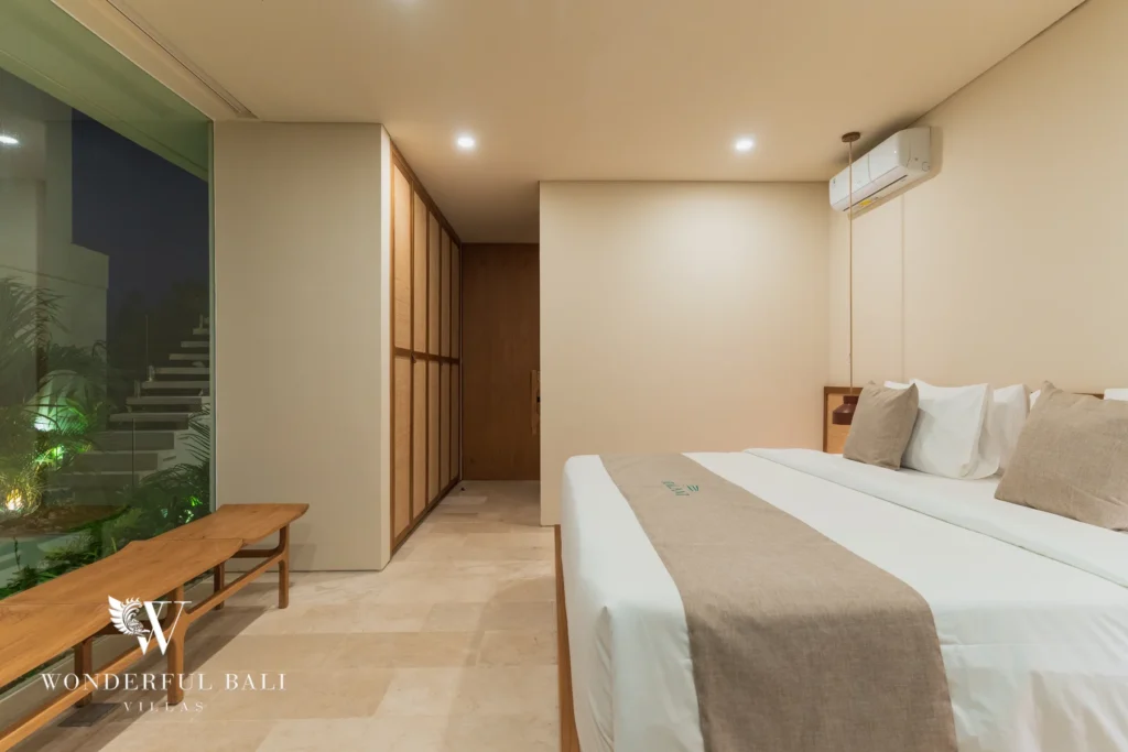 A bedroom at night with a double bed, timber bench and a glass wall overlooking lit jungle greenery at a 3-bedroom villa in Bingin, Uluwatu, Bali