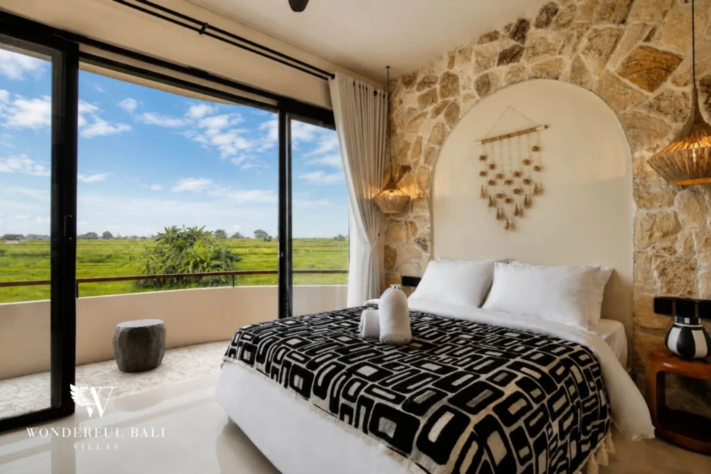 A queen bedroom with a stone feature wall and balcony opening to rice fields at a 3-bedroom villa in Pererenan, Bali