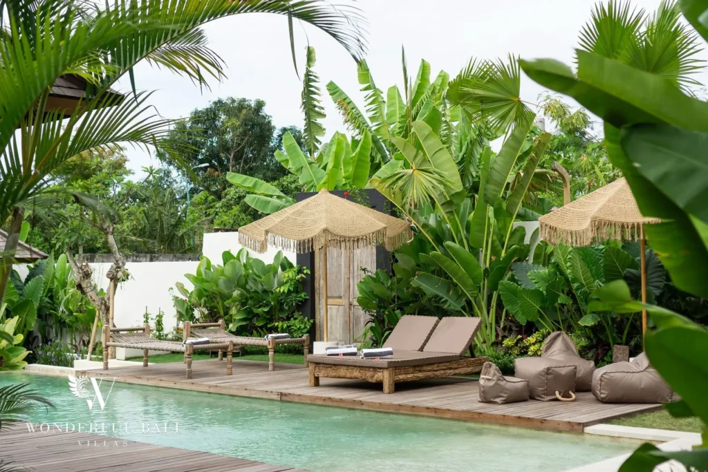 A poolside deck with rattan sun loungers, fringed umbrellas and bean bags is framed by palms and banana plants beside the pool at a 4-bedroom villa in Canggu, Bali