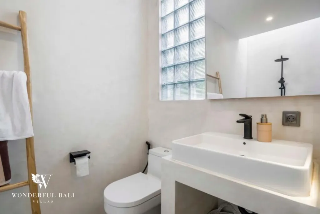A micro-cement bathroom with a white vessel sink, black tapware, glass-block window, and timber towel ladder at a 3-bedroom villa in Pererenan, Bali